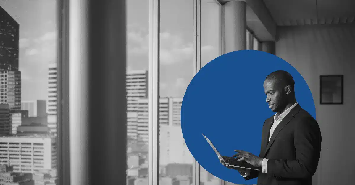 A man looks out a window at a city skyline while checking email on a laptop.