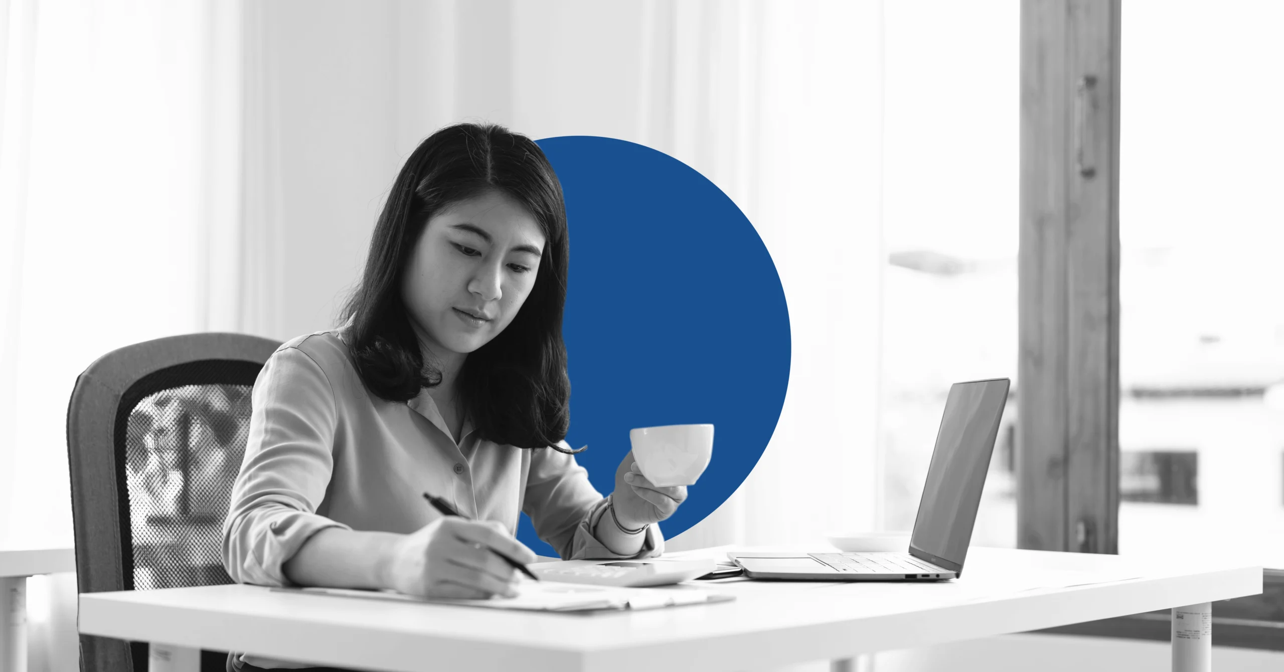 A businesswoman drinks from a cup and takes notes while working on a laptop.